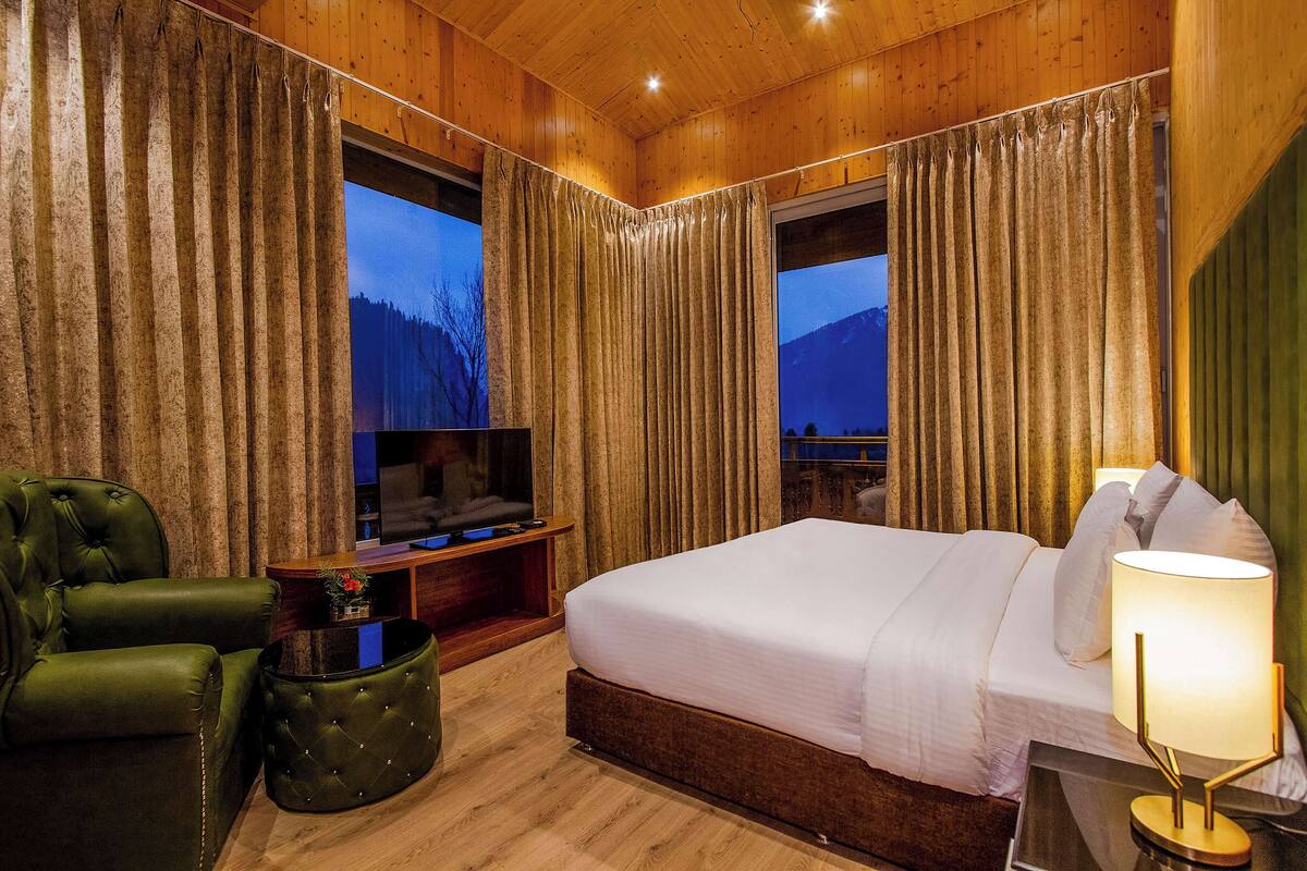 Una habitación de hotel con una cama grande, un sillón verde, un televisor y vistas a las montañas desde las ventanas.