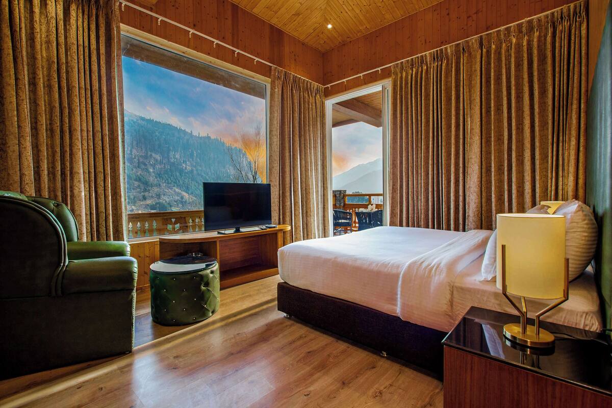 Una habitación de hotel con un ventanal grande, una cama, un televisor, una lámpara y un sillón verde.