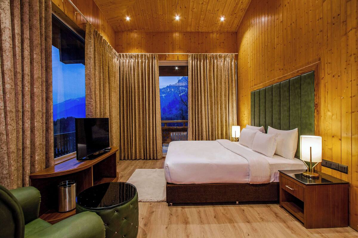 Habitación de hotel con interior de madera, una cama con sábanas blancas, un televisor sobre un mueble de madera, un sillón verde y vistas a montañas a través de las cortinas.