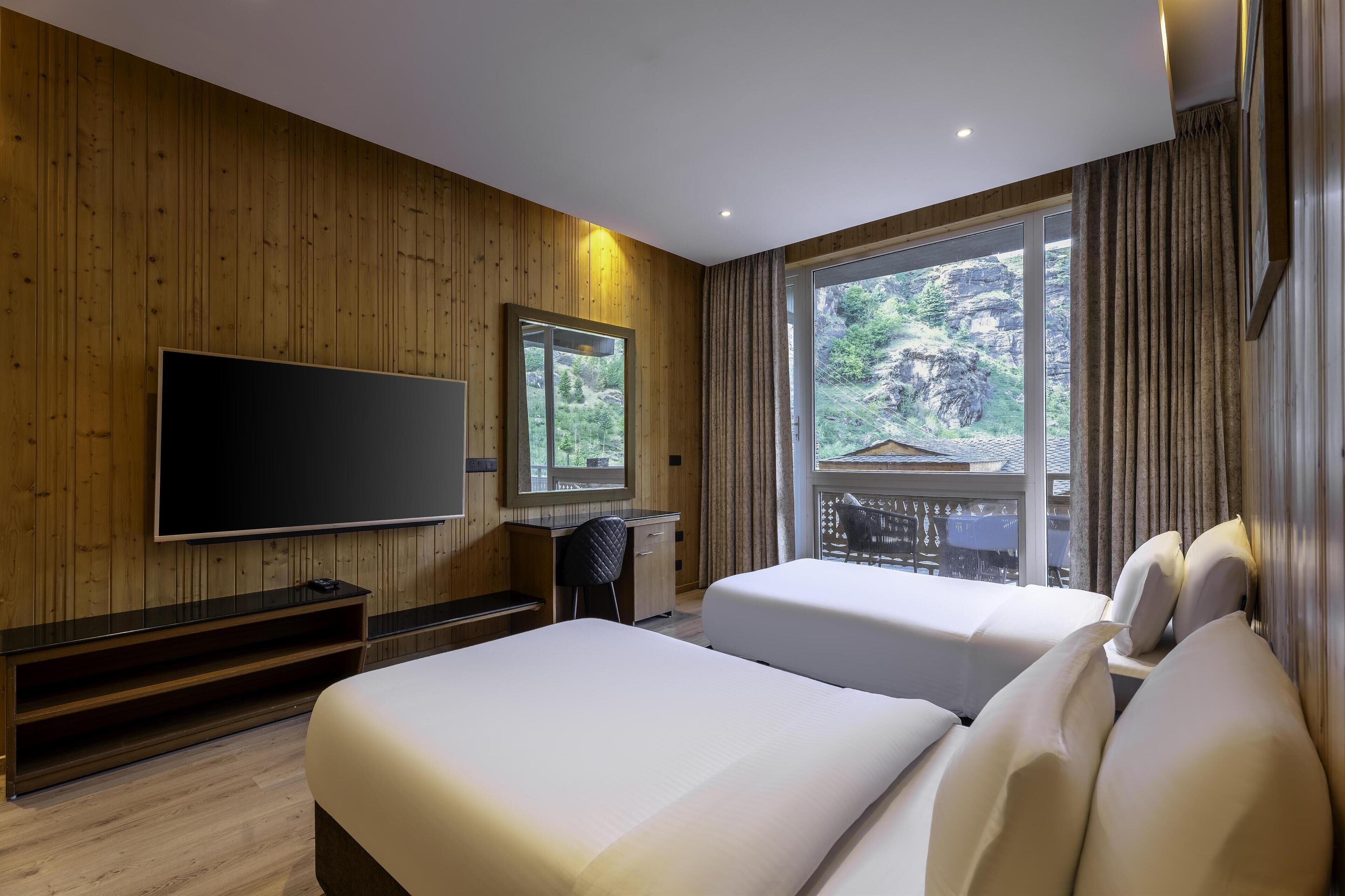 A hotel room with wooden walls, a large TV, two beds, a desk, and a window with a view of greenery.