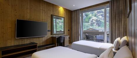 A hotel room with wooden walls, a large TV, two beds, a desk, and a window with a view of greenery.