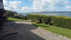 Vista de un paisaje costero con un área cubierta de hierba, un sendero pavimentado y una masa de agua al fondo.