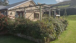 Una casa de piedra con una pérgola de madera, rodeada de abundante vegetación y un cielo despejado.