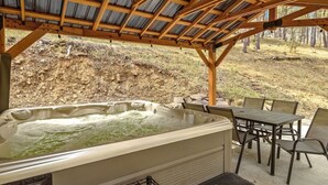 Uma banheira de hidromassagem sob uma pérgola de madeira, com uma mesa e cadeiras, em meio a uma paisagem natural.