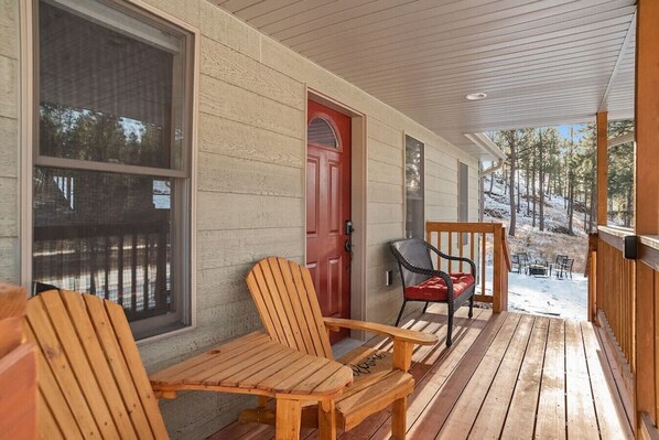 Une véranda en bois avec deux fauteuils Adirondack et un fauteuil à bascule, une porte rouge et une vue sur un paysage enneigé.
