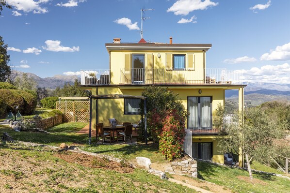 A two-story house with a balcony, a patio with outdoor furniture, and a view of mountains in the background. - House Girasole in the country with Pool, garden and spectacular sea view (Toscana)