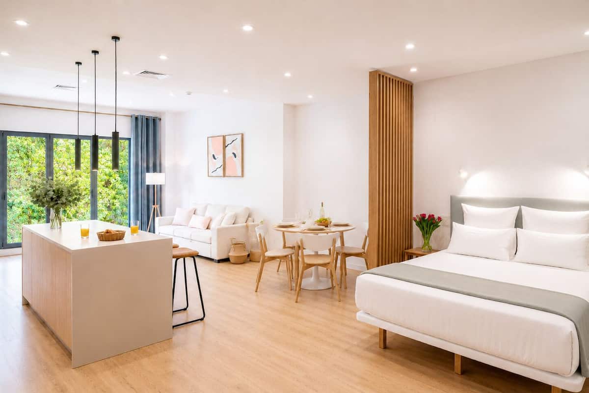 A modern hotel room with a bed, a dining area, a kitchenette, and large windows.