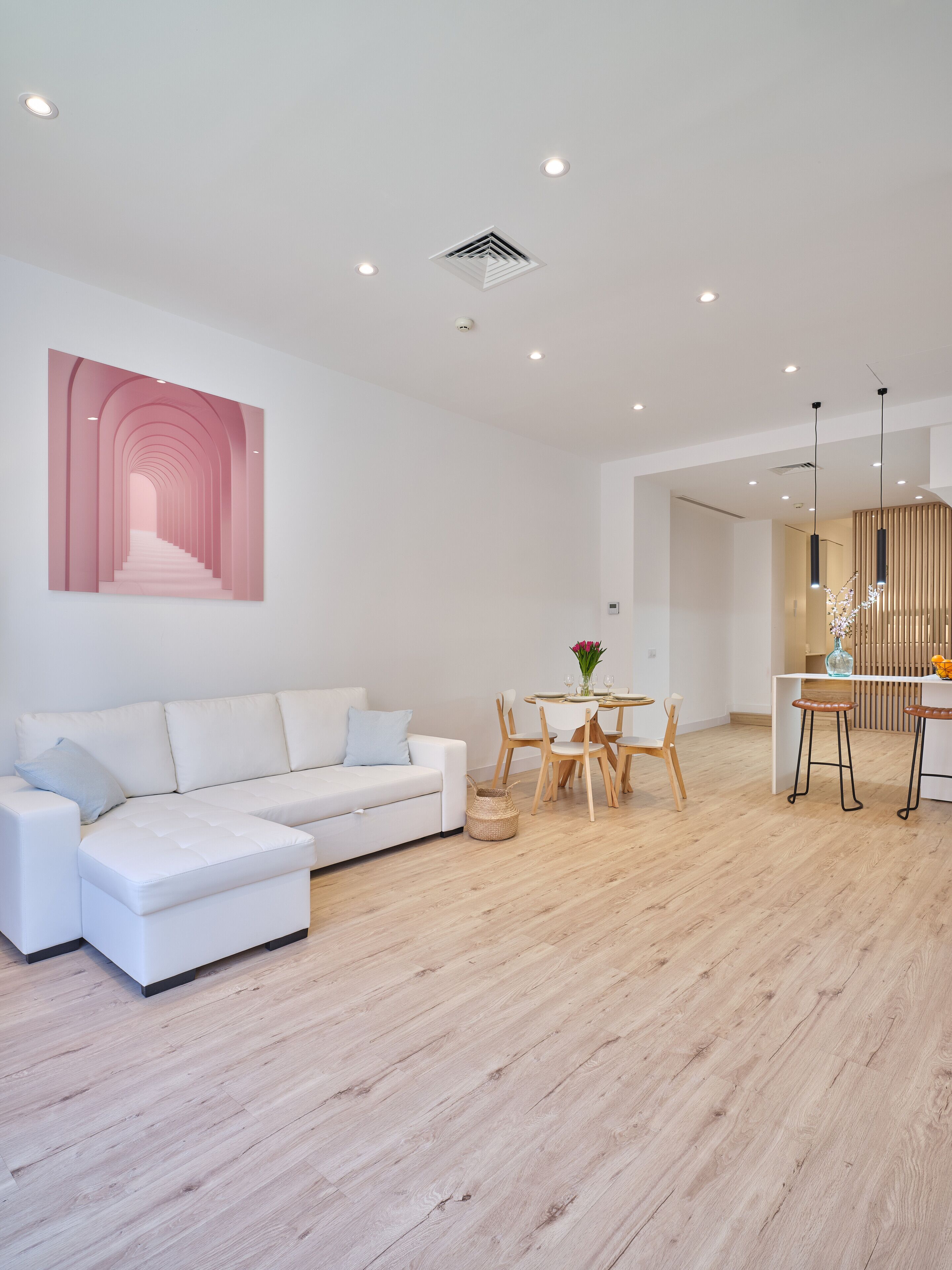 Una sala de estar moderna con un sofá seccional blanco, un comedor con una mesa redonda y sillas, y una cocina con armarios de madera y un lámpara colgante.