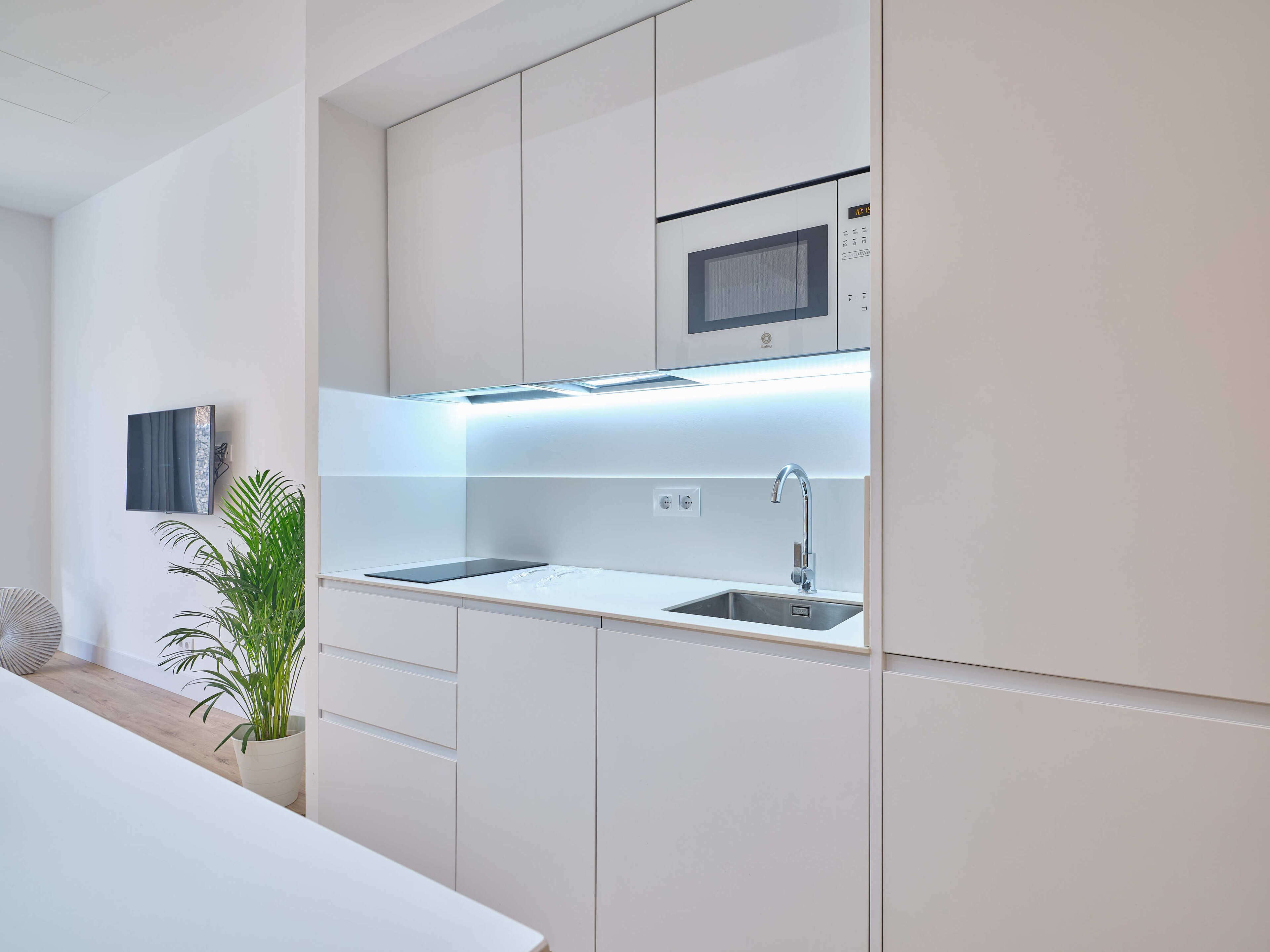 A modern kitchen with white cabinetry, a built-in microwave, a stainless steel sink, and under-cabinet lighting.