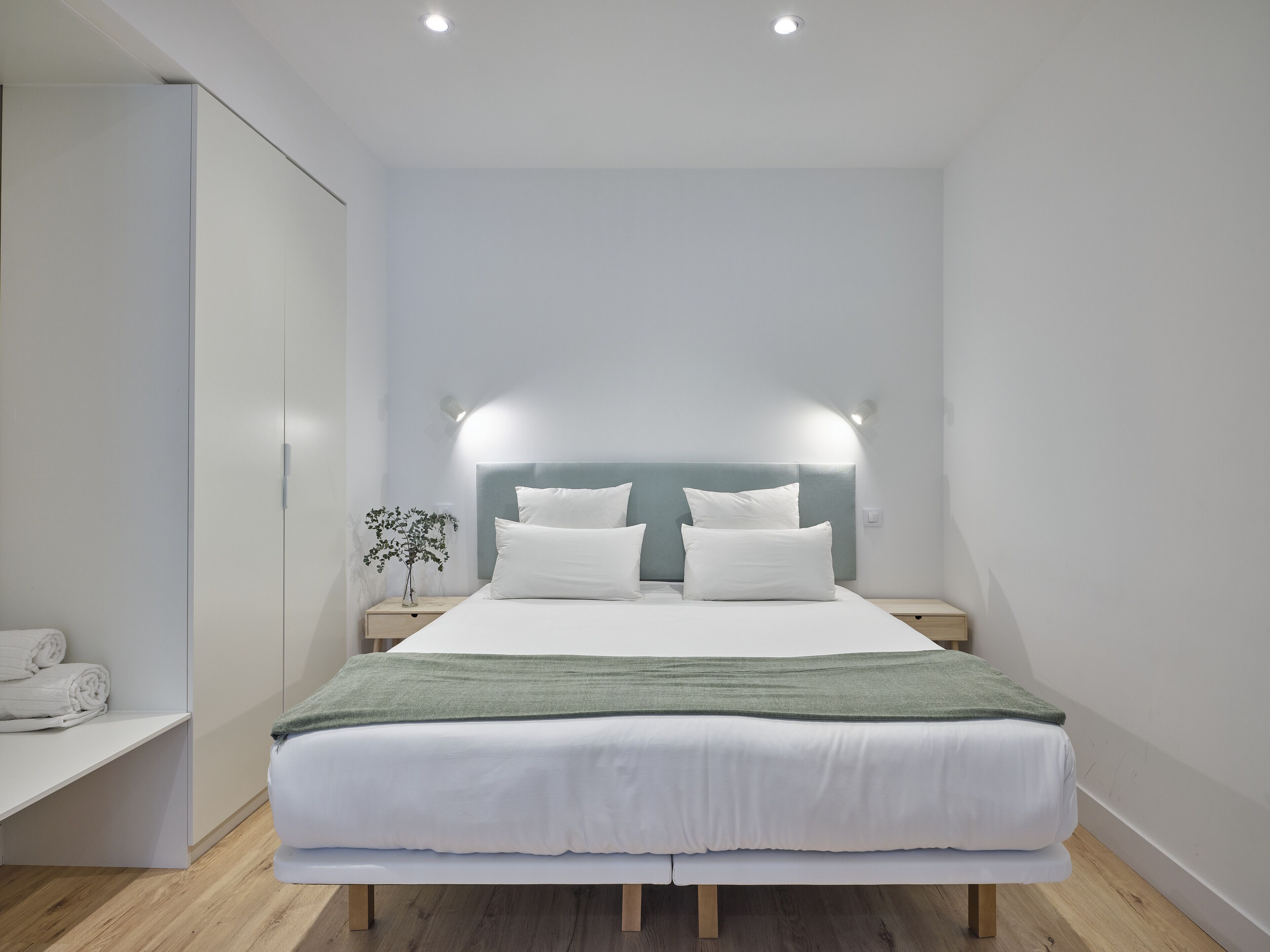 A modern bedroom with a large bed, two bedside tables, a nightstand, a wall-mounted light, and a small plant.