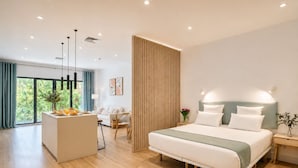 Ein modernes Hotelzimmer mit einem großen Bett, einer Sitzecke mit Tisch und Stühlen, einer Küchenarbeitsplatte und einem großen Fenster mit Vorhängen.