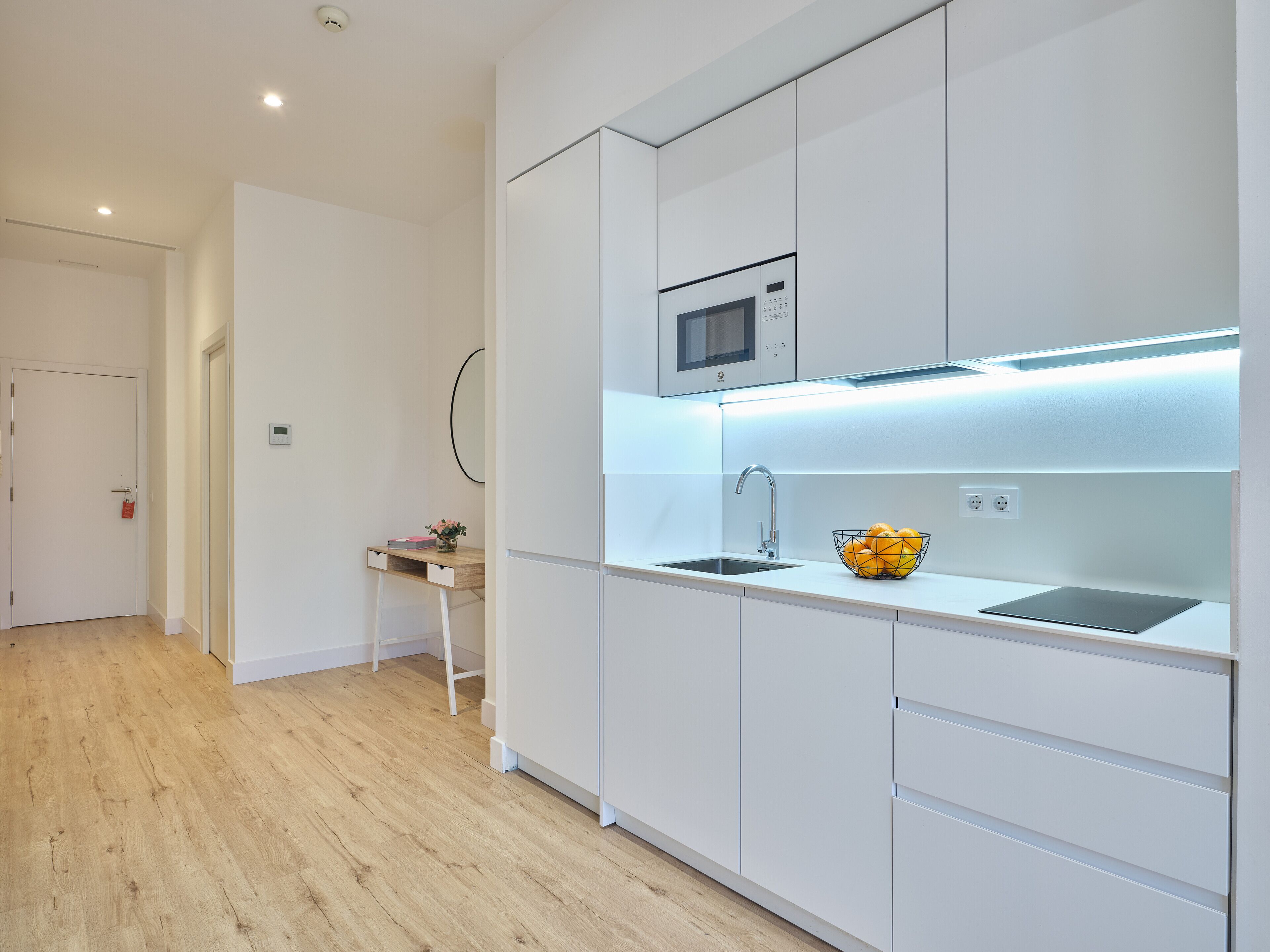 Una cucina moderna con mobili bianchi, un forno a microonde integrato, un lavandino e una ciotola di frutta sul piano di lavoro.
