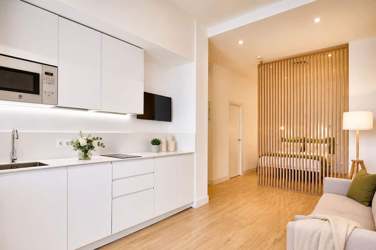 A modern kitchen with white cabinetry, a built-in microwave, and a sink. There is a dining area with a wooden divider and a sofa.