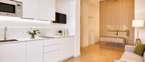 A modern kitchen with white cabinetry, a built-in microwave, and a sink. There is a dining area with a wooden divider and a sofa.