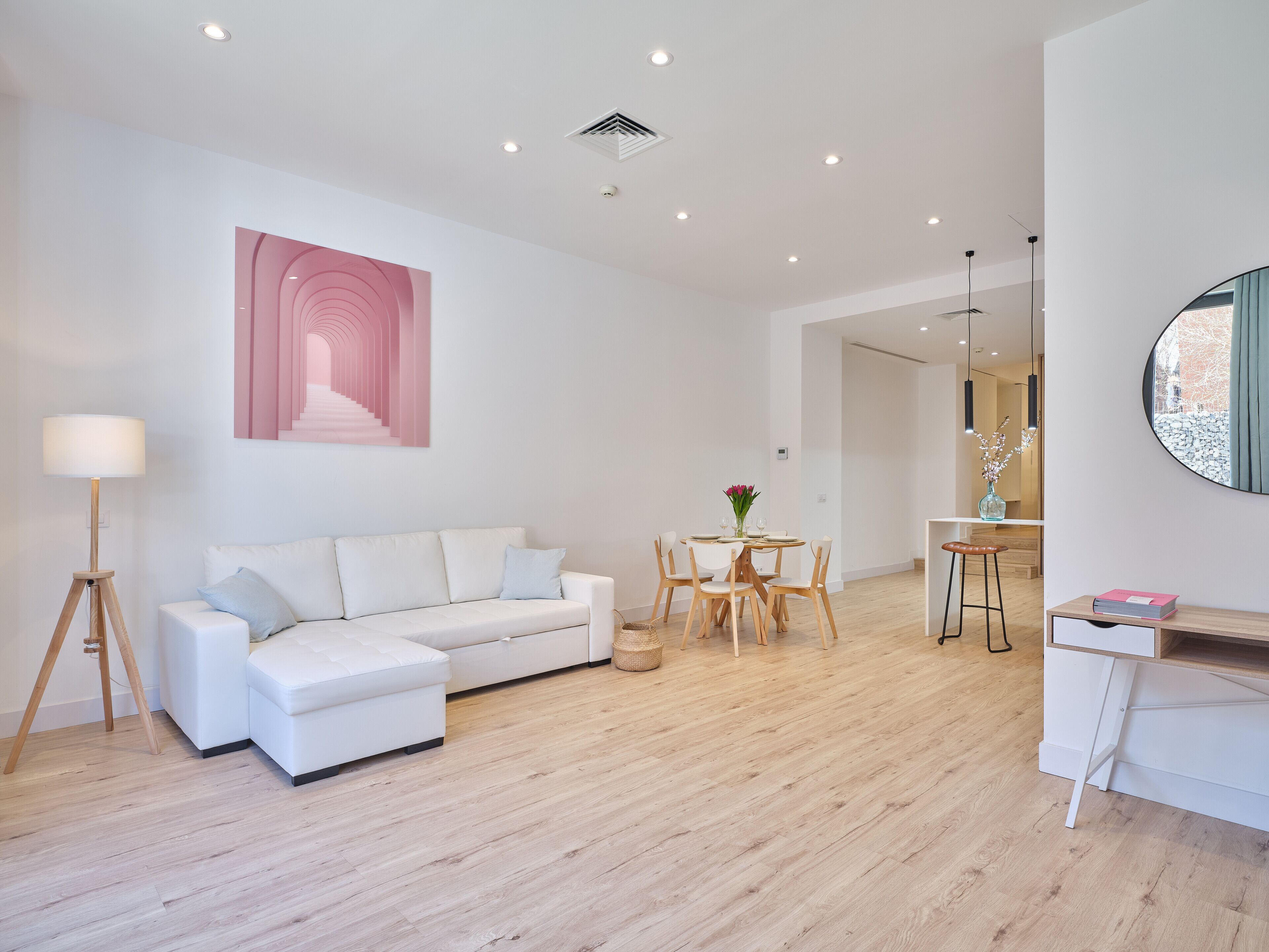 A modern living room with a white sofa, a dining table with chairs, and a small kitchen area with a stool.