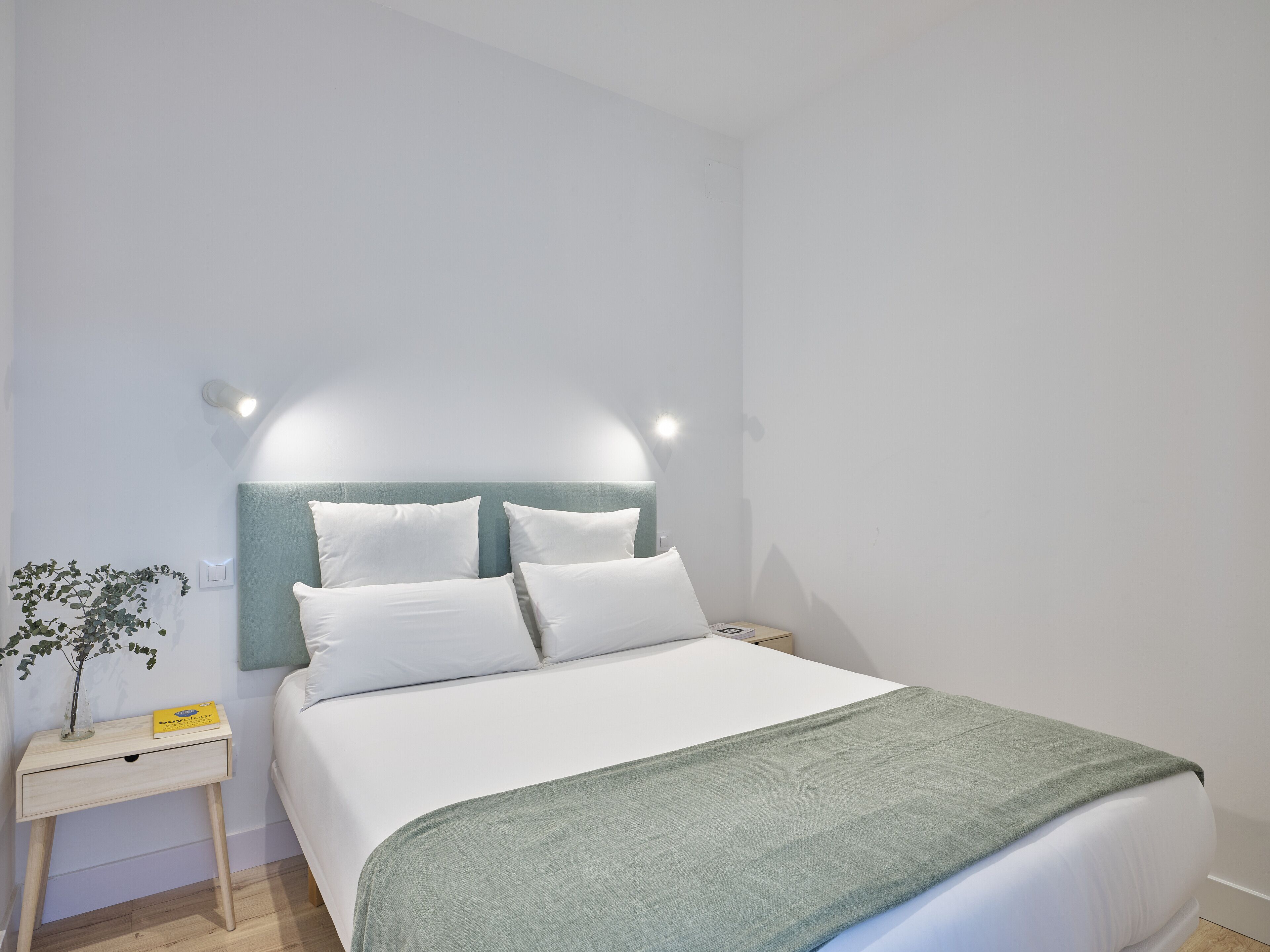 Una cama bien hecha con sábanas blancas y un plaid verde, una mesita de noche con lámpara y una planta pequeña.