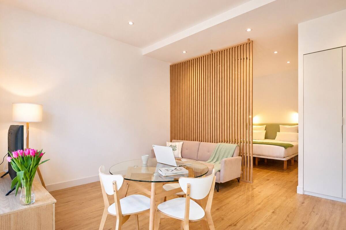 A modern living room with a glass dining table, white chairs, a sofa, and a bed.