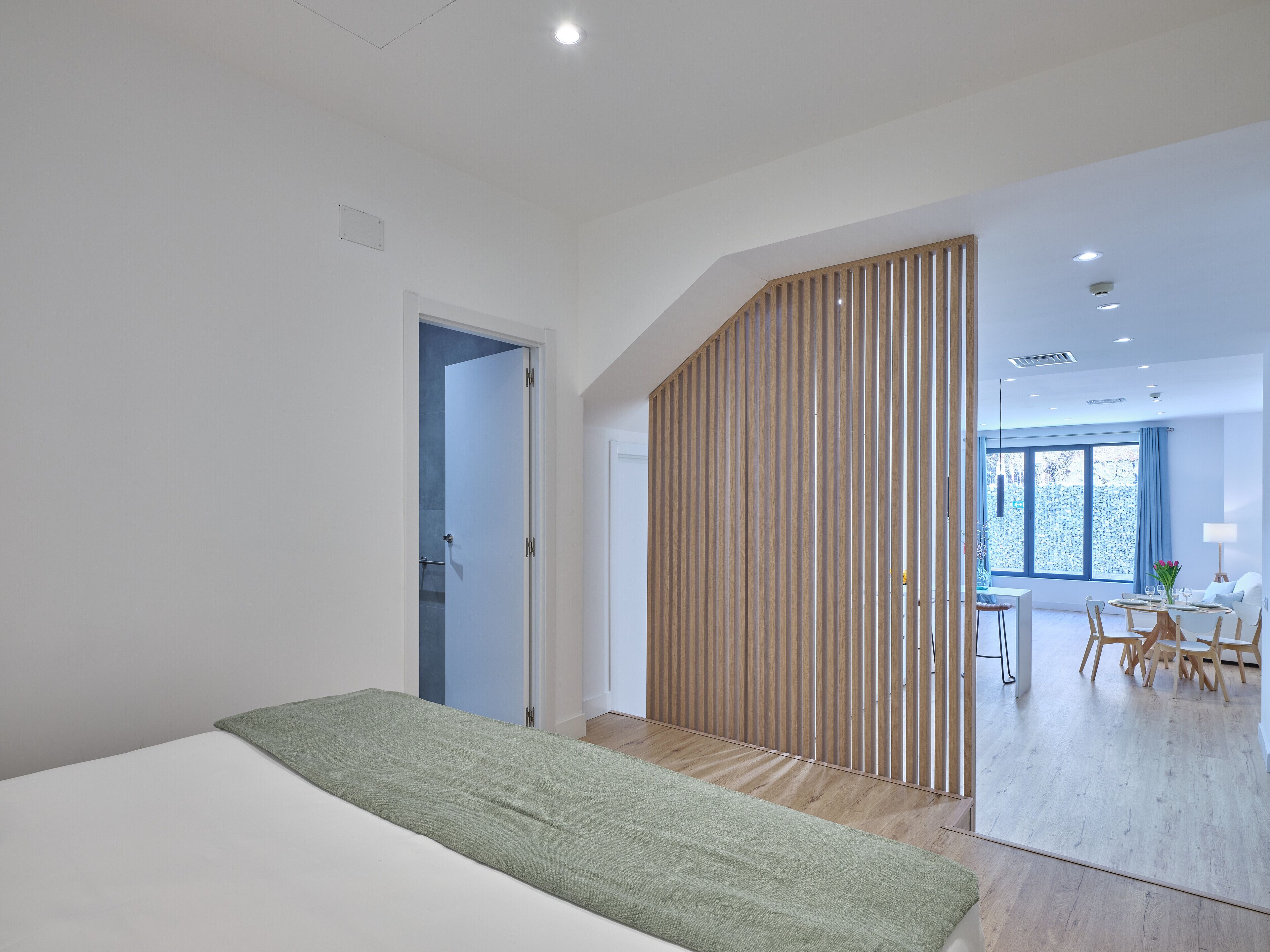 A modern hotel room with a bed, a wooden partition, and a dining area visible through an open door.