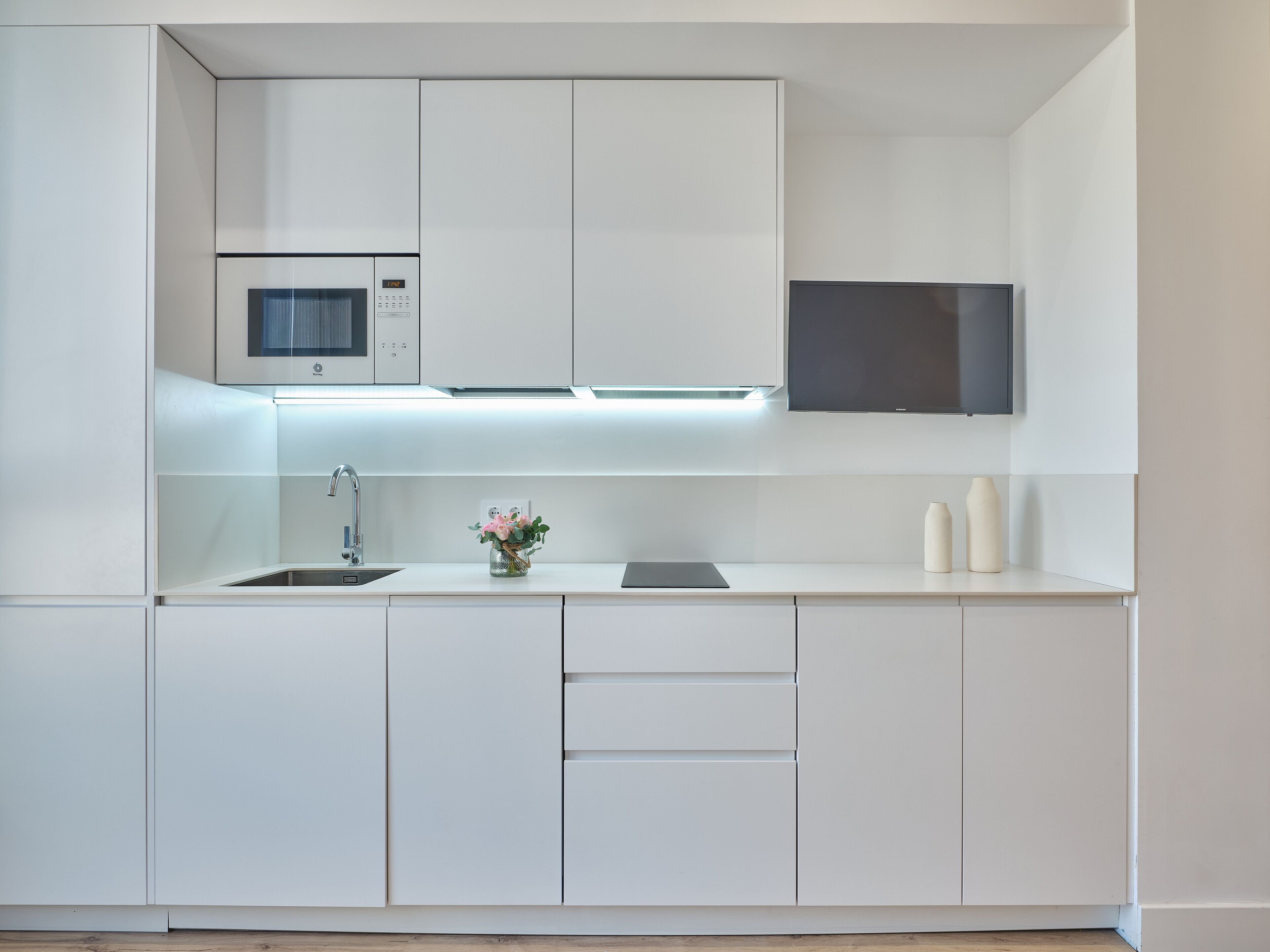 Una cocina moderna con armarios blancos, un microondas, un lavamanos y un televisor de pantalla plana sobre el mostrador.