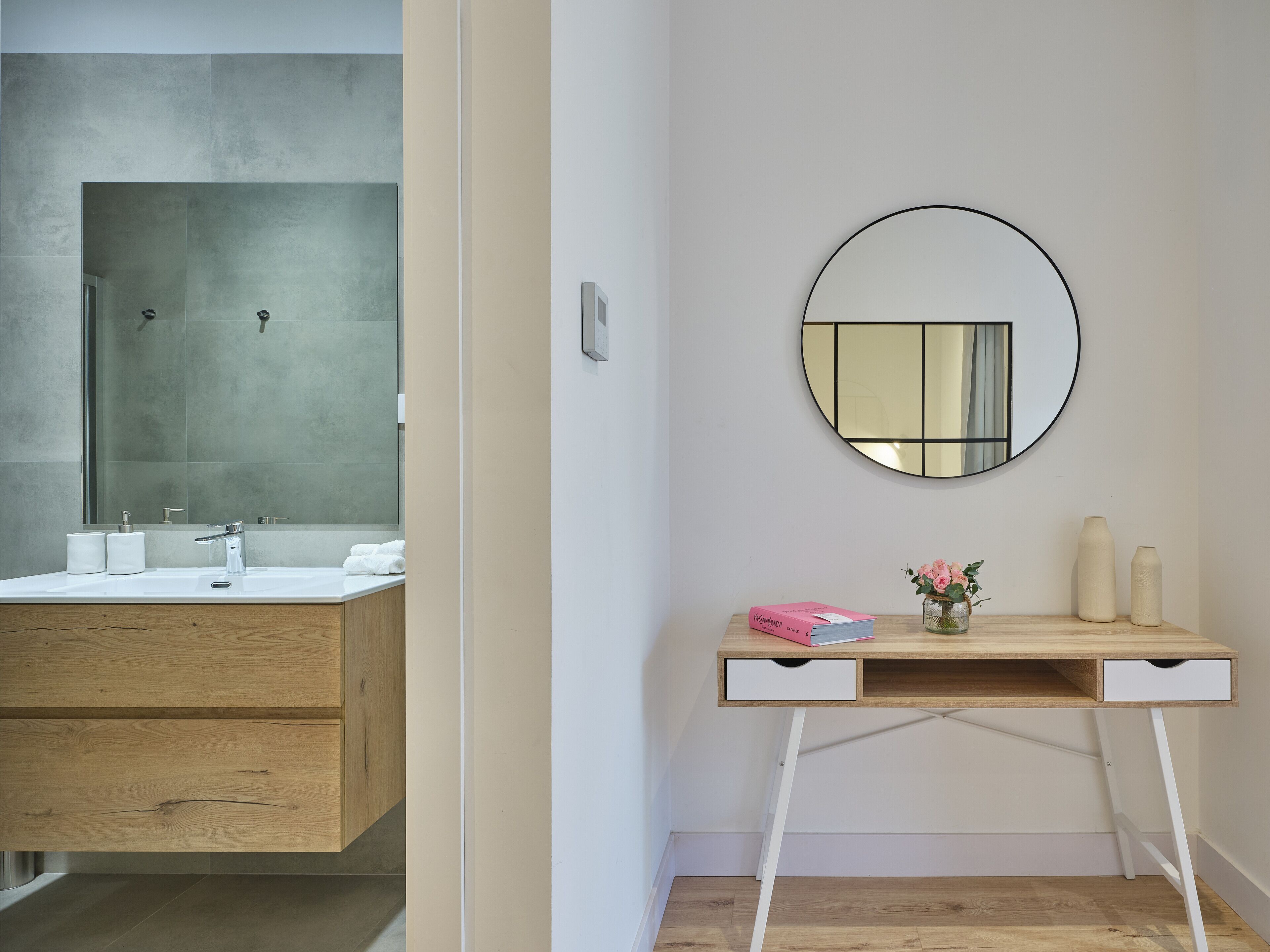 A modern bathroom with a large mirror, a wooden vanity, and a small desk with a vase and books.