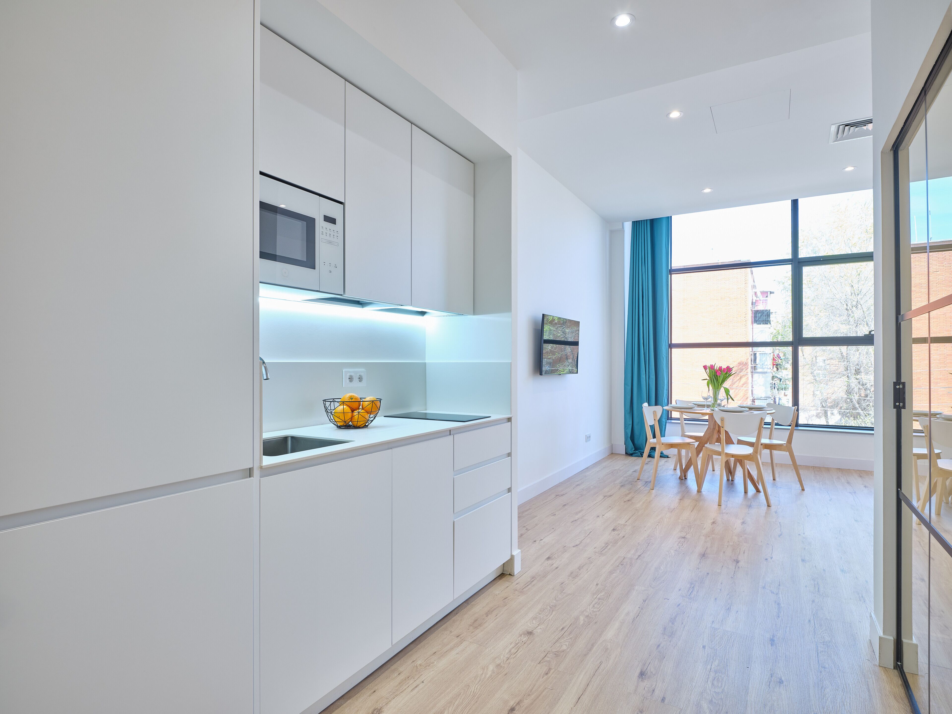 A modern kitchen with white cabinets, a microwave, and a fruit bowl. There is a dining area with a table and chairs, and a large window with curtains.