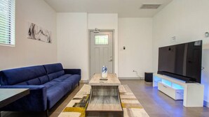 A modern living room with a blue sofa, a coffee table with a vase, a flat-screen TV, and a door leading to another room.