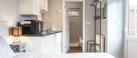 Un dormitorio moderno con cama, una kitchenette pequeña y un baño visible a través de una puerta abierta.