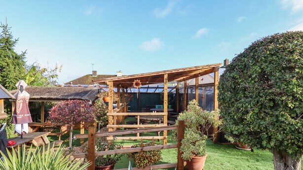 A wooden pergola with a glass enclosure, surrounded by potted plants and a well-maintained garden.