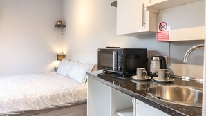 A compact hotel room with a bed, a kitchenette with a microwave, a coffee maker, and a sink. There is a 'No smoking' sign above the sink.