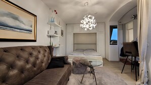 Ein Wohnzimmer mit braunem Ledersofa, einem Glastisch, einem Bett mit weißer Bettwäsche, einem weißen Kleiderschrank und einem einzigartigen Kronleuchter.