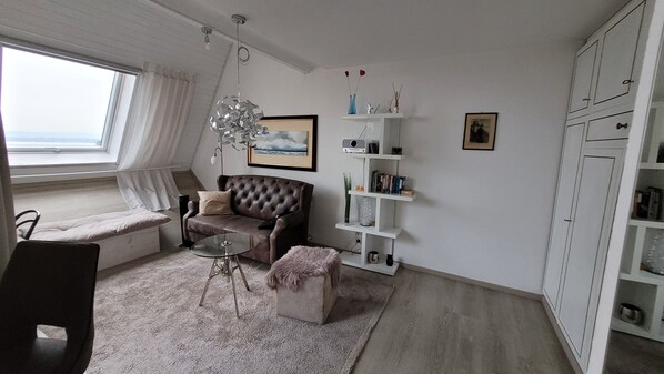 A living room with a sofa, a glass coffee table, a small ottoman, a white bookshelf, and a window with curtains.