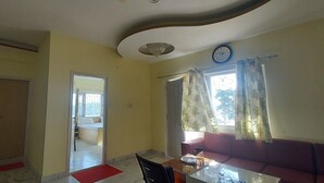 A living room with a sofa, a dining table, a red mat, a clock, and a window with curtains. - LakeBreeze By Sunrise (Nainital)
