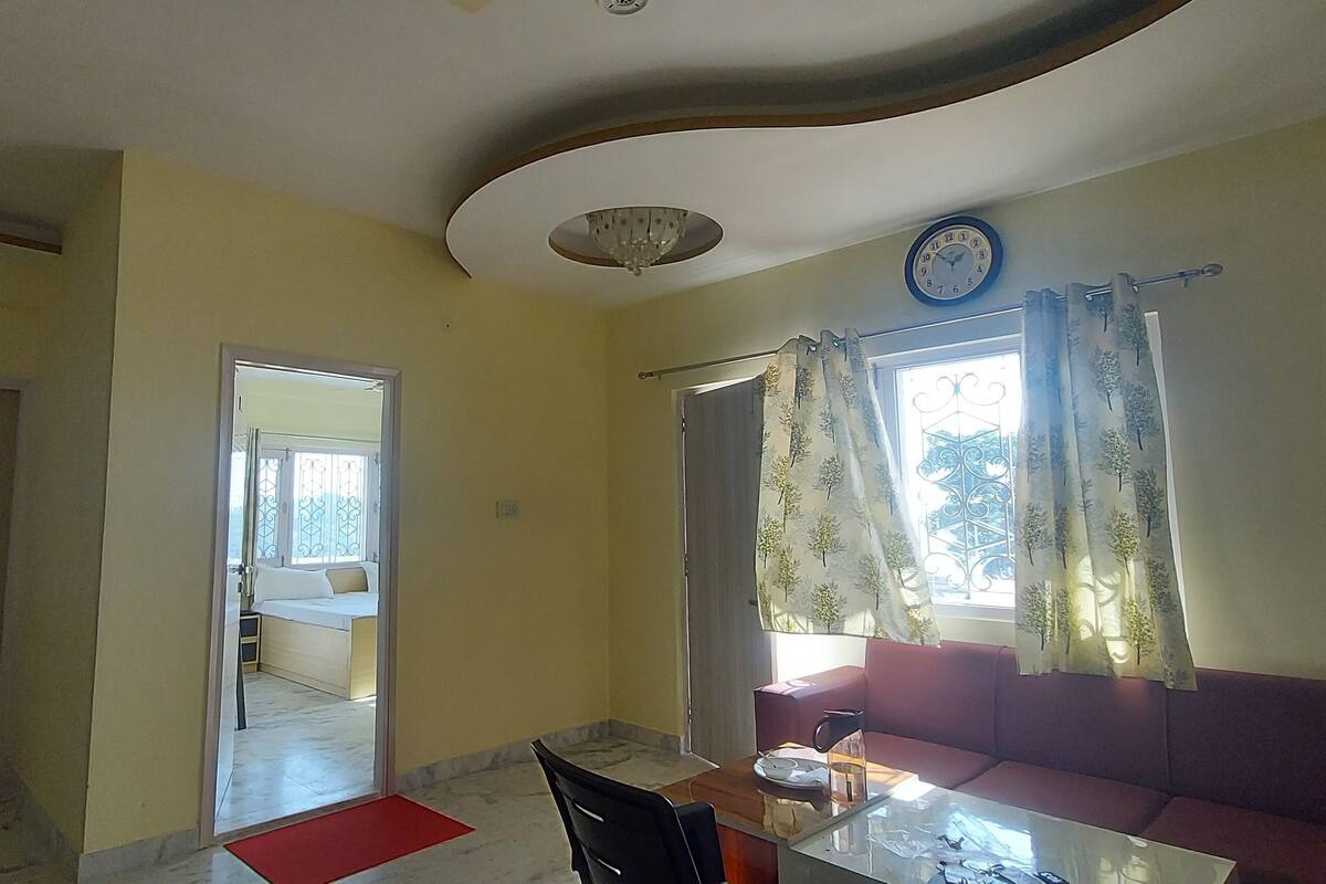 A living room with a sofa, a dining table, a red mat, a clock, and a window with curtains.