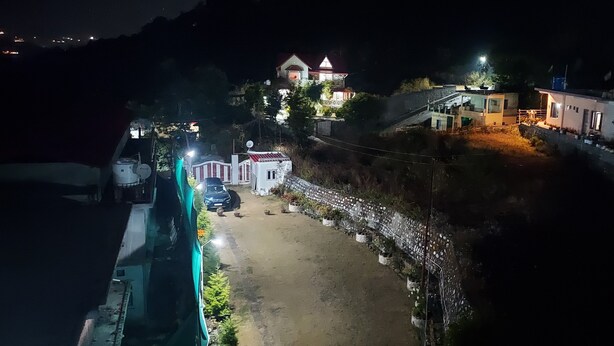 A nighttime view of a residential area with houses, a paved road, and a small white building with a red roof. - LakeBreeze By Sunrise (Nainital)