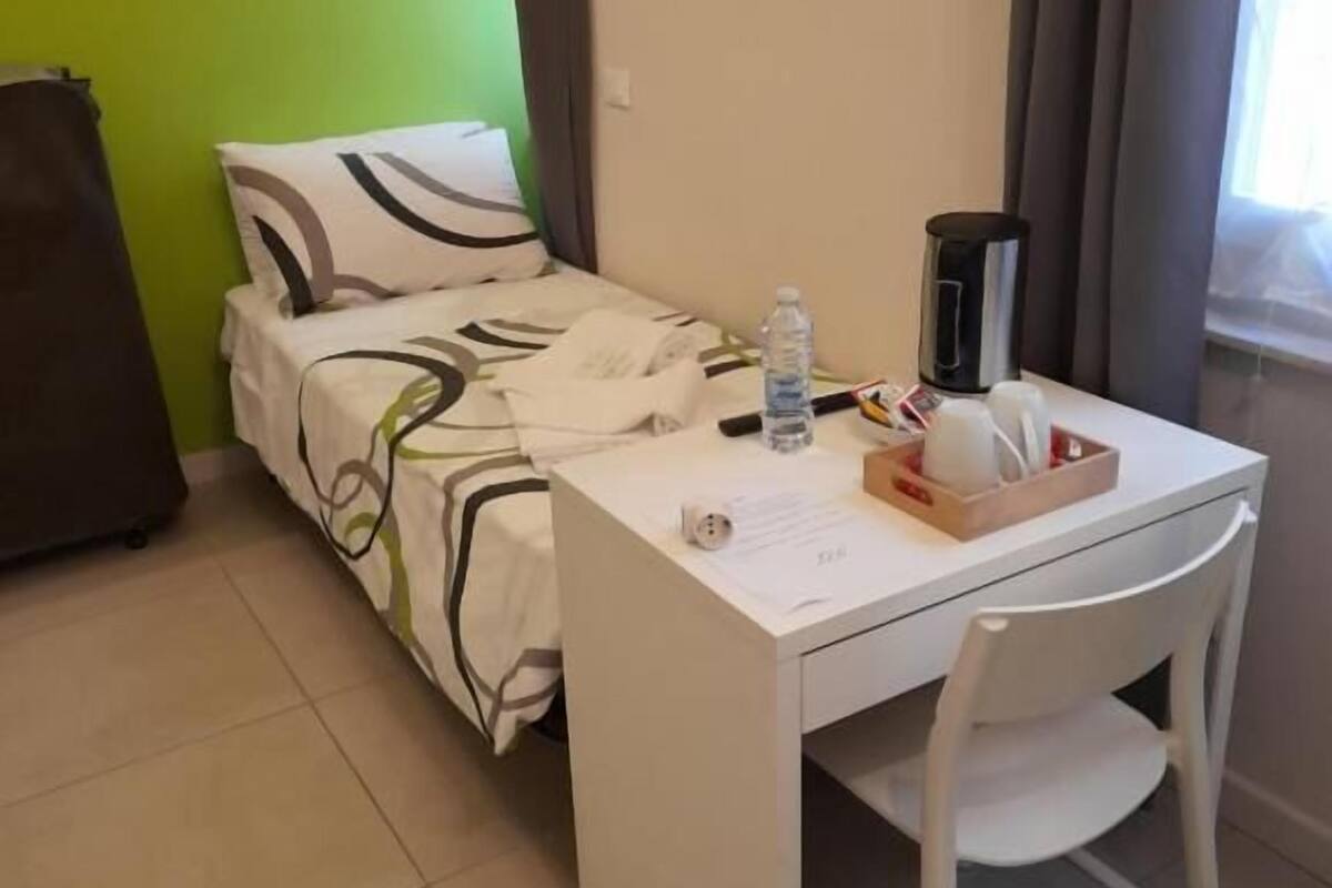 Een kleine, verzorgde hotelkamer met een bed, een bureau en een stoel. Op het bureau staat een waterfles, een beker en een dienblad met een theepot.
