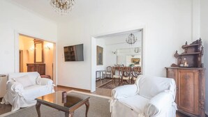 A living room with two white armchairs, a glass coffee table, a wooden cabinet, and a dining area in the background. - Classic Athenean Apartment at Acropolis Museum & Metro (Athens)