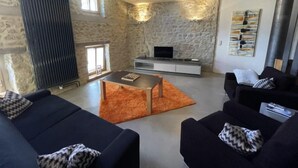 Un salon moderne avec des murs en pierre, une cheminée, un téléviseur à écran plat et deux canapés noirs ornés de coussins à motifs.