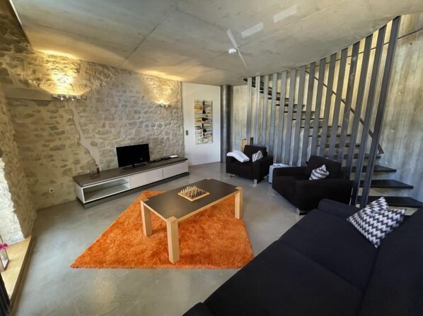A modern living room with a stone wall, a flat-screen TV, a wooden coffee table, a black sofa, and a staircase with metal railings.