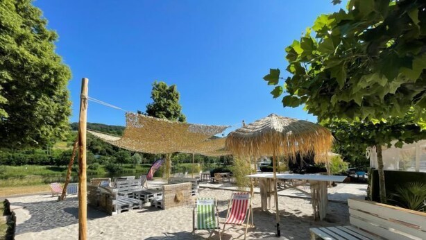Un espace aménagé au bord de la plage, avec des bancs en bois, un parasol en paille et un drapeau.