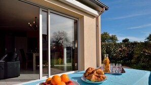 An outdoor dining area with a table set for breakfast, including fruits and pastries, and a bottle of juice. - Charme de Penvins by Interhome (Sarzeau)