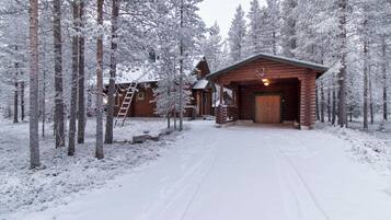 A log cabin with a wooden porch and a ladder leading to a loft, surrounded by snow-covered trees.