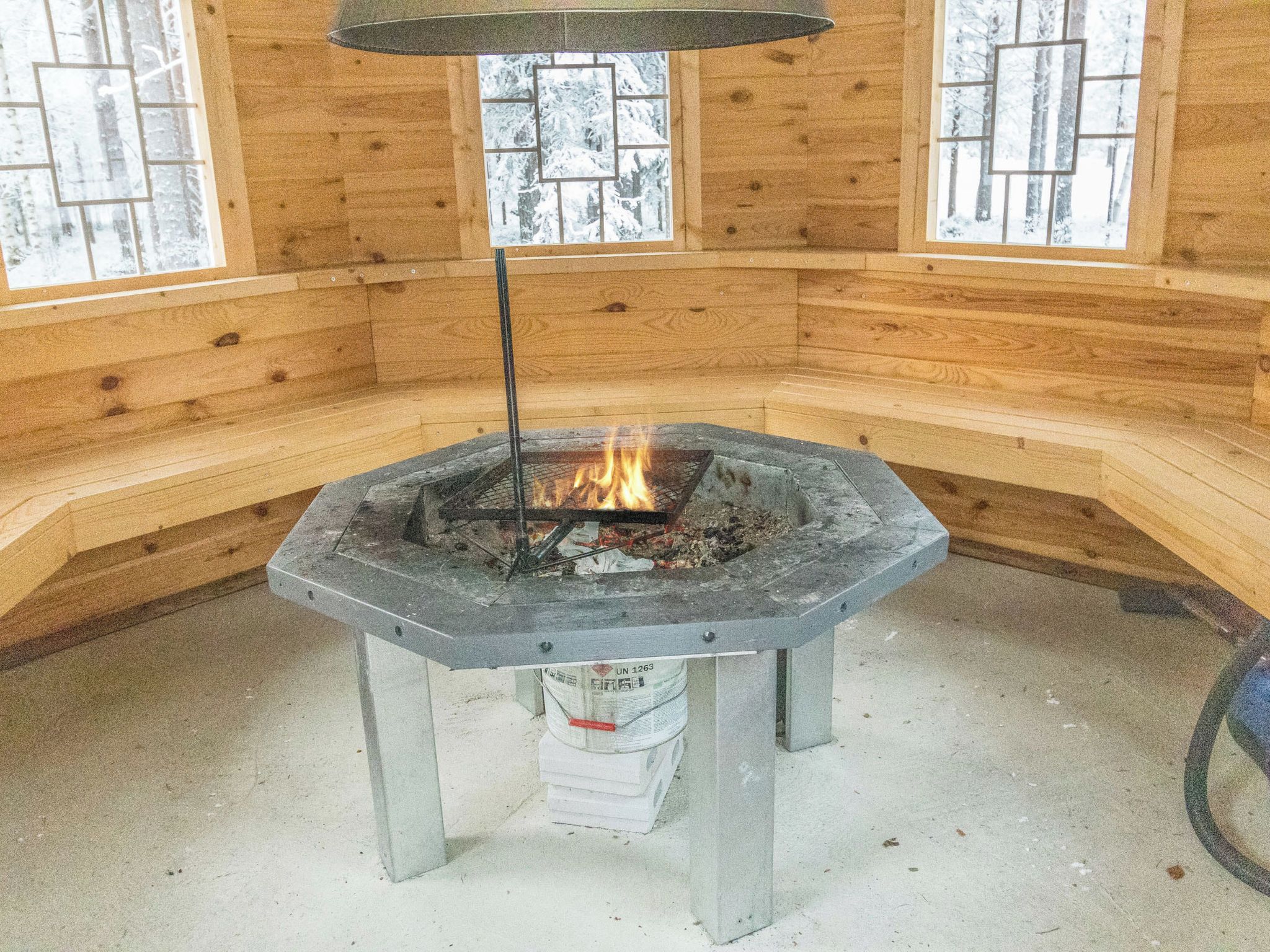 Une pièce aux murs en bois et dotée d’un foyer central en pierre où l’on peut voir les flammes.