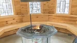 Een vuurkuil van steen met een metalen standaard in een houten hut met grote ramen.