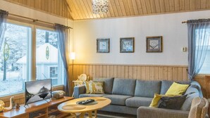 Een gezellige woonkamer met een bank, fauteuil en een salontafel. Drie ingekaderde foto’s hangen aan de muur en er is een raam met gordijnen.