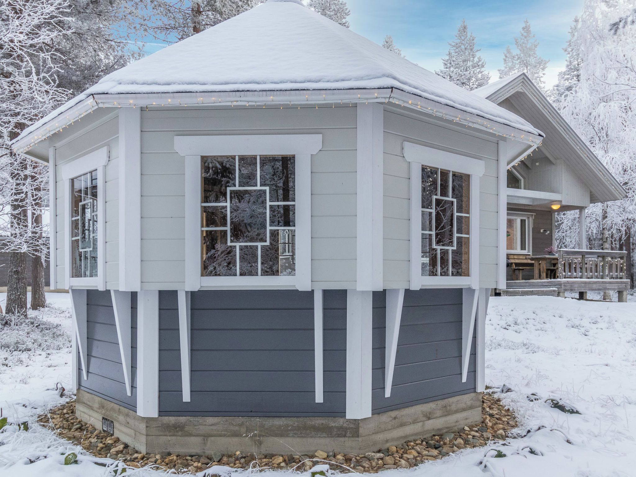 A snow-covered gazebo with a dark siding exterior and white trim.