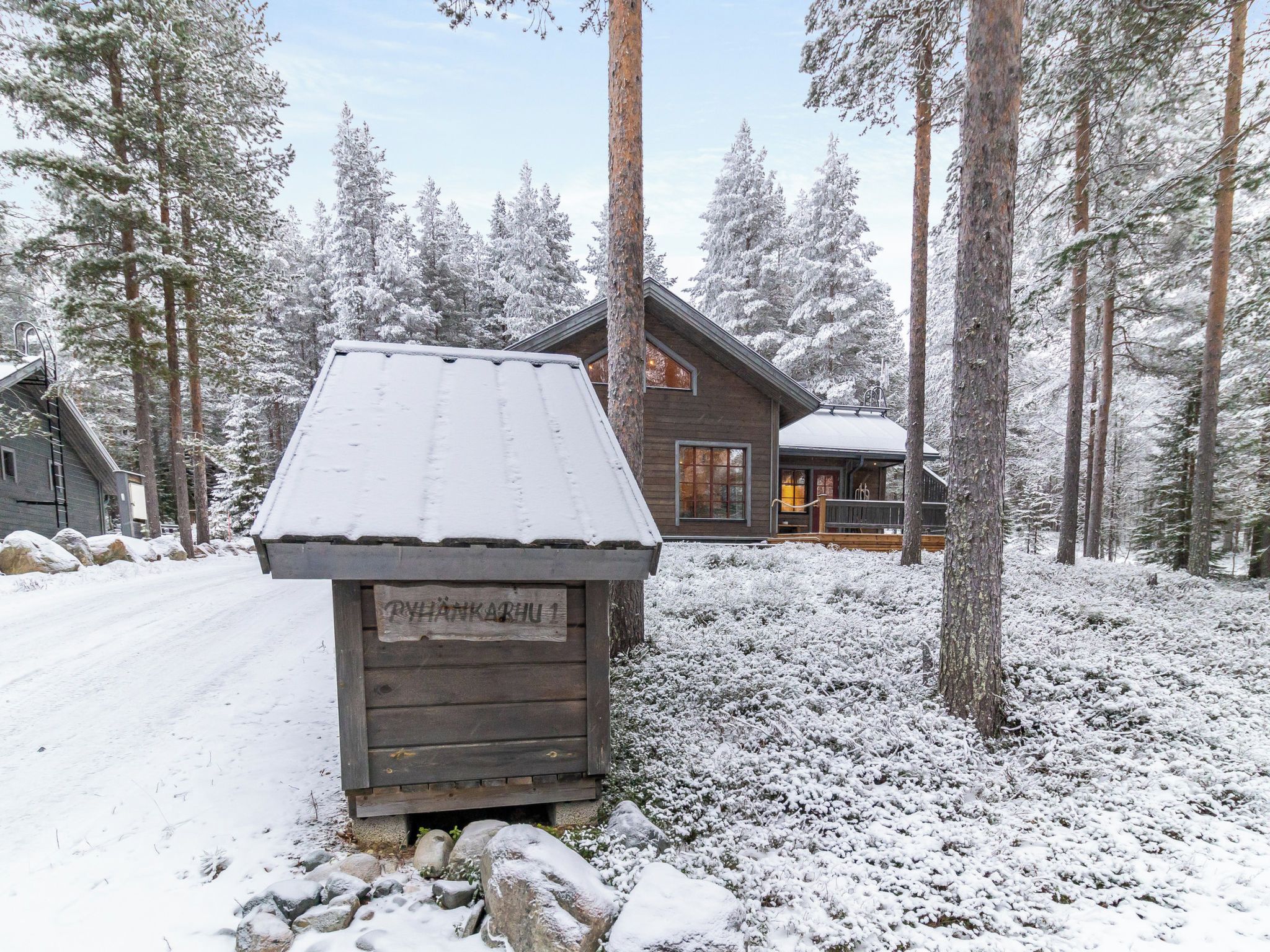 Lumihunnun peittämä puinen mökki, jonka ympärillä kohoaa korkeita mäntyjä.