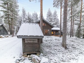 Lumihunnun peittämä puinen mökki, jonka ympärillä kohoaa korkeita mäntyjä.