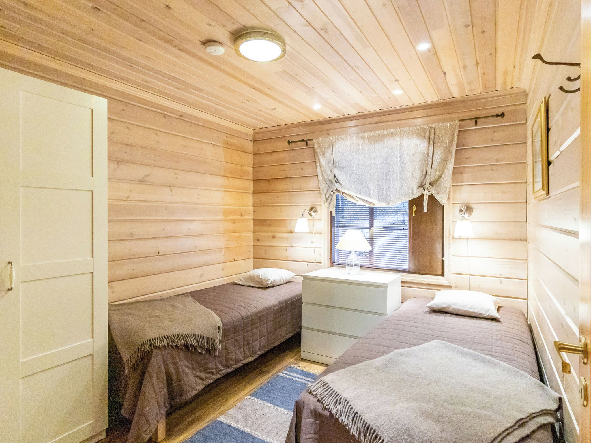 A wooden cabin room with two beds, a desk, and a window with curtains.