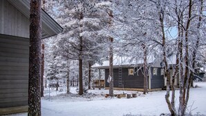 Una cabaña con terraza de madera y una escalera que lleva al techo, rodeada de árboles cubiertos de nieve y un edificio de madera.