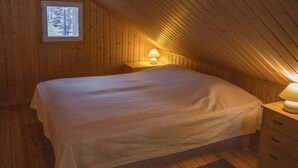 Un dormitorio acogedor en el ático, con techo inclinado de madera, una cama individual, una mesita de noche con lámpara y un ventanal con vistas a árboles.